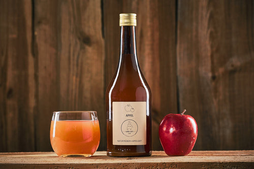 Das Bild zeigt Land.Luft Apfelsaft, präsentiert zwischen einem Apfel und einem Glas Apfelsaft. Der naturbelassene, aromatische Saft eignet sich perfekt als Erfrischung.