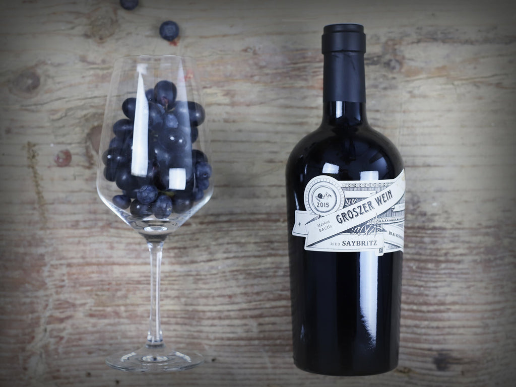 Das Bild zeigt eine Flasche Groszer Wein Saybritz neben einem dekorativen Weinglas mit Weintrauben. Die elegante Präsentation unterstreicht die Qualität des Weins und eignet sich perfekt für Weinliebhaber, besondere Anlässe oder als Geschenk.